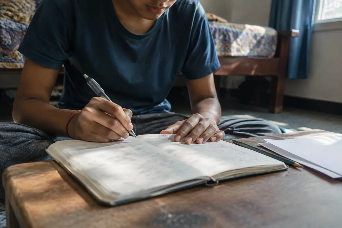 A student sitting on the floor beside a low table, pen in hand over an open paper diary, daylight falling across the page