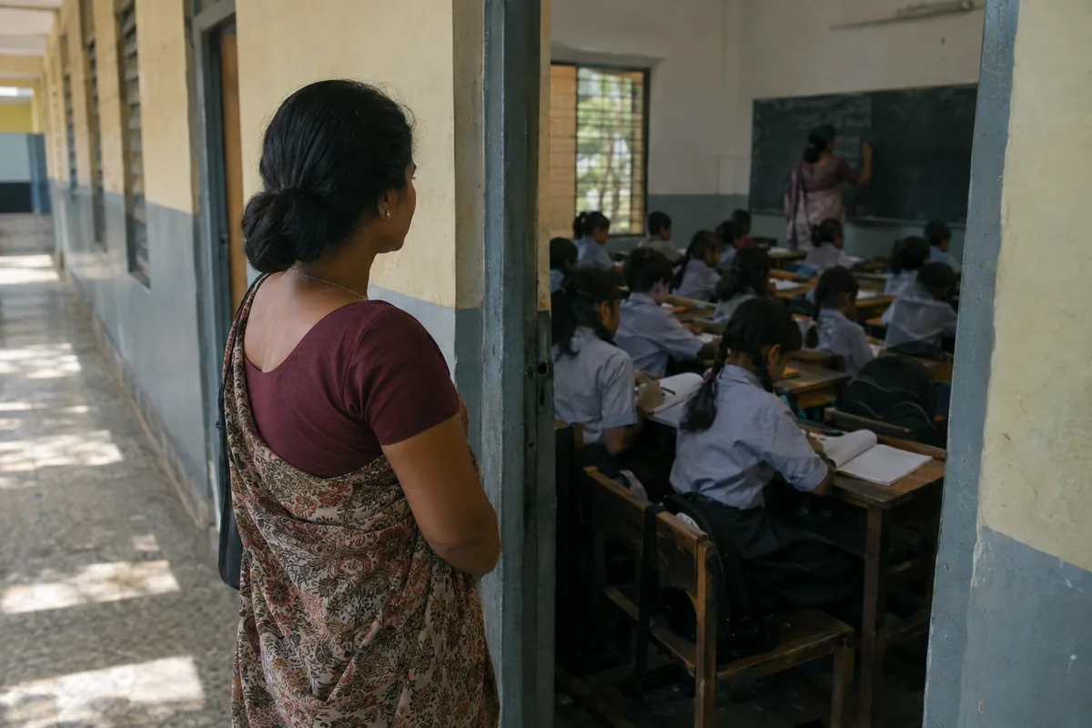 A parent standing at an open classroom doorway in a school corridor, looking in at children in uniform writing at rows of wooden desks while a teacher works at the blackboard