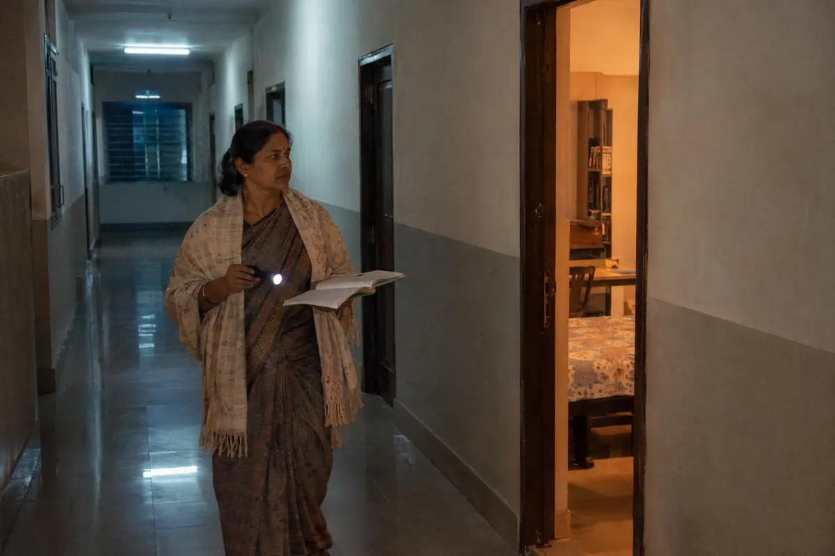 A hostel warden in a saree and shawl walks a dim corridor at night with a small torch and an open register in her hand, pausing by an open door of a lit student room