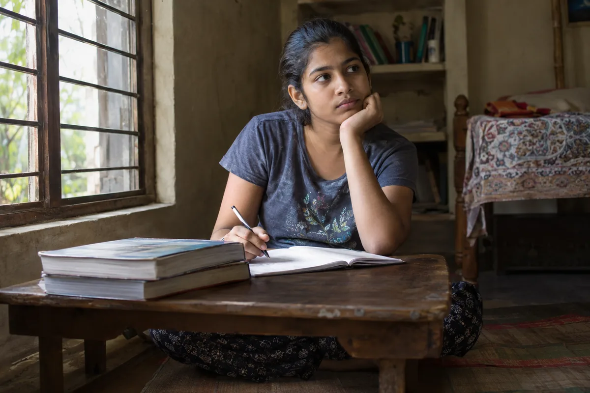 A student revising alone at a low table in a home study corner, textbooks stacked and a notebook open in front of them