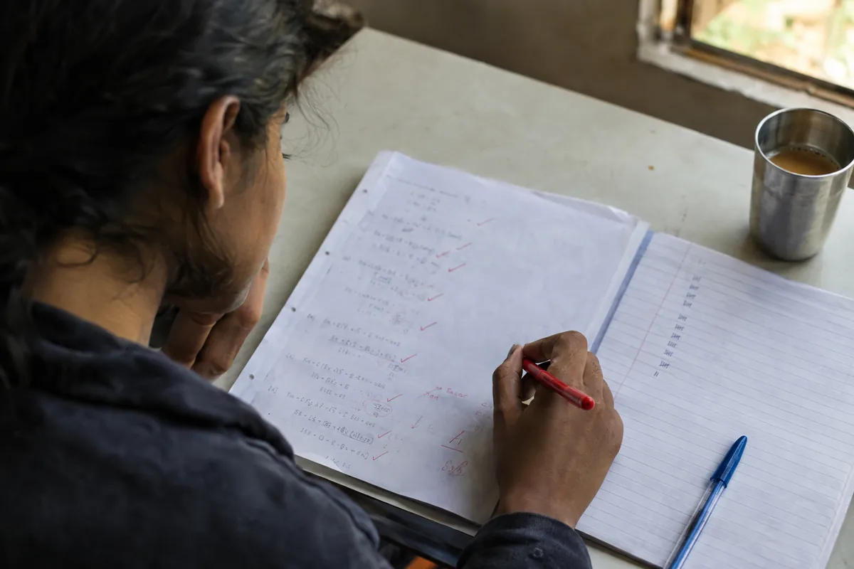A student marking a corrected answer script with a red pen, a notebook with a short tally of pen strokes beside it